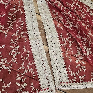 Op de afbeelding: Een dieprode, doorschijnende sjaal of omslagdoek met ingewikkeld goudkleurig bloemenborduursel over het gehele materiaal. De randen zijn afgewerkt met een brede, geschulpte gouden kanten bies die langs de lengte van de stof loopt.