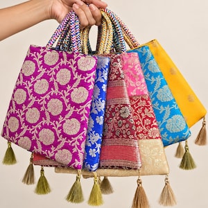 Op de afbeelding: Een collectie rechthoekige stoffen tasjes met ingewikkelde bloemen- en geometrische patronen in levendige kleuren zoals magenta, blauw, rood en geel. Elk tasje heeft gevlochten handvatten en een decoratief kwastje aan de onderste hoek.