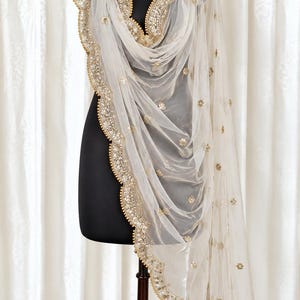Op de afbeelding: Een transparante, gebroken witte dupatta-sjaal met een geschulpte rand voorzien van ingewikkeld goudborduurwerk, metalen lovertjes en kleine kraaltjes. De stof is versierd met verspreide gouden bloemmotieven over de gehele lengte.
