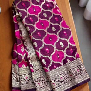 May include: A folded textile with a repeating pattern of magenta and purple ovals, accented with gold and white floral designs. The border features a detailed floral pattern with gold and white accents. The fabric is draped on a wooden surface.