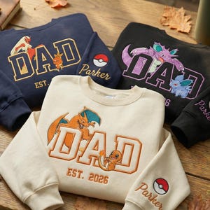 May include: Navy, black, and cream crewneck sweatshirts. Each has the word "DAD" in a collegiate font, with Pokemon characters and the name "Parker" on the sleeve. One sweatshirt has "EST. 2026".