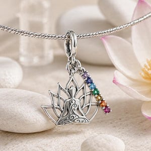 Puede incluir: Colgante de plata con un diseño de figura meditando dentro de una flor de loto. Una barra con gemas de colores cuelga del dije. El colgante está suspendido de una cadena de plata, sobre un fondo suave y neutro.