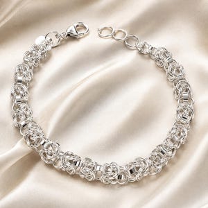 Könnte beinhalten: Ein silbernes Kettenarmband mit einem komplexen byzantinischen Flechtmuster. Das Armband besteht aus miteinander verbundenen Metallgliedern und verfügt über einen Karabinerverschluss sowie einen kleinen runden Anhänger am Verschluss.