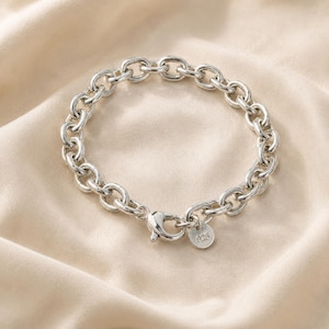 Op de afbeelding: Een zilverkleurige schakelarmband met een karabijnsluiting en een klein, rond label. De armband bestaat uit ovale schakels. De achtergrond is een lichtbeige stof.