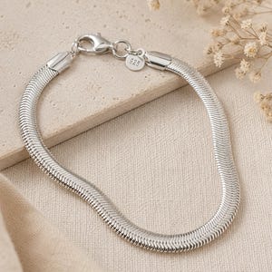 Könnte beinhalten: Ein silberfarbenes Schlangenkettchen-Armband mit polierter Oberfläche und Karabinerverschluss. Ein kleiner, runder Metallanhänger ist in der Nähe des Verschlusses befestigt und deutlich mit der Zahl 925 graviert, was das Sterlingsilber des Schmucks anzeigt.