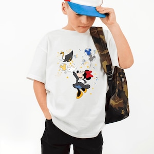 Peut inclure: T-shirt blanc avec un motif coloré de Minnie Mouse attrapant des ballons. Le motif comprend un cygne noir, des ballons en forme de tête de Mickey Mouse et des feux d'artifice. Le t-shirt est porté par une personne avec un sac à dos et une casquette de baseball bleue et blanche.