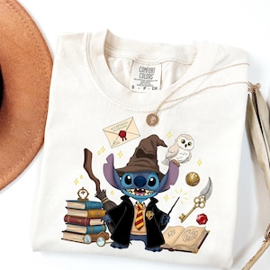 May include: Cream-colored t-shirt featuring a cartoon Stitch character dressed as a wizard, complete with a hat, robe, and wand. The design includes a stack of books, an owl, and other magical elements. The shirt has the Comfort Colors brand label.
