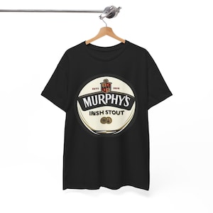 Op de afbeelding: Zwart T-shirt met een cirkelvormig logo voor Murphy's Irish Stout. Het logo is wit en goudkleurig met de woorden "Murphy's Irish Stout" en een wapen. Het T-shirt hangt aan een houten kleerhanger.
