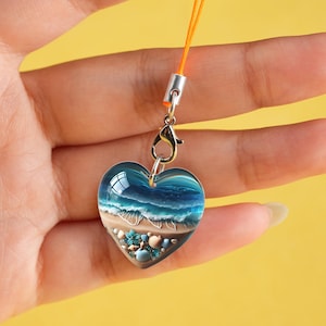 May include: Heart-shaped charm with a beach scene. The clear resin charm features a blue ocean with white waves, a sandy shore, and seashells. It has a silver clasp and an orange lanyard.