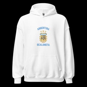 Puede incluir: Una sudadera con capucha blanca que presenta un gráfico central con el texto ARGENTINA en azul claro, tres estrellas doradas y el escudo de la AFA en oro y azul. Debajo del escudo, el texto SCALONETA está impreso en azul claro en el pecho.