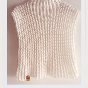Puede incluir: Un top de crochet blanco sin mangas con cuello alto, colgado de una percha blanca. El top tiene una textura acanalada y una pequeña etiqueta marrón. Las palabras "Chaleco Blanco" están escritas en cursiva en la parte inferior.
