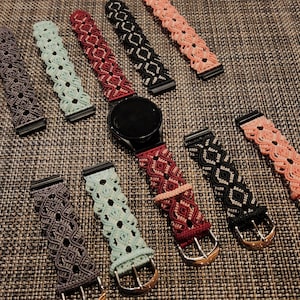 May include: A black smartwatch with several macrame watch bands in various colors, including gray, teal, red, black, and peach. The bands feature intricate woven patterns and metal hardware for attachment. The watch bands are approximately 0.75 inches wide.
