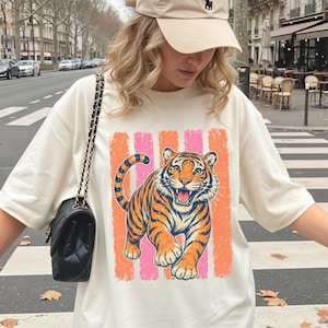 May include: A beige baseball cap with a small dark logo and an oversized cream-colored t-shirt featuring a graphic of a tiger in orange and black stripes against a background of vertical pink and orange textured stripes, paired with a black quilted bag.