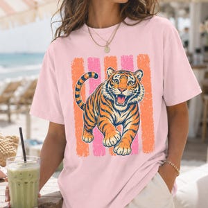 May include: A light pink short-sleeve t-shirt featuring a graphic print of a roaring tiger. The tiger is orange with black stripes and is positioned in front of a background of vertical, distressed-texture stripes in alternating orange and pink.