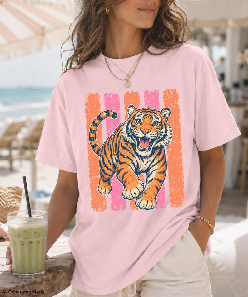 May include: A light pink short-sleeve t-shirt featuring a graphic print of a roaring tiger. The tiger is orange with black stripes and is positioned in front of a background of vertical, distressed-texture stripes in alternating orange and pink.
