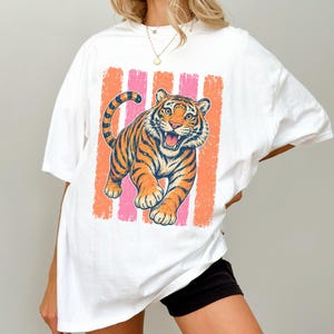 May include: A white short-sleeved t-shirt featuring a graphic print of a tiger with orange and black stripes. The tiger is positioned in front of vertical stripes in shades of coral and pink. The shirt has a crew neckline and a loose fit.