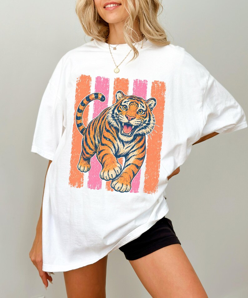 May include: A white short-sleeved t-shirt featuring a graphic print of a tiger with orange and black stripes. The tiger is positioned in front of vertical stripes in shades of coral and pink. The shirt has a crew neckline and a loose fit.