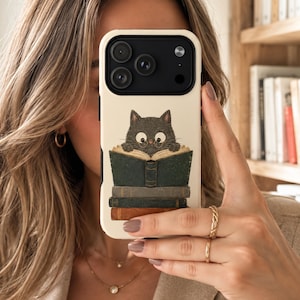 May include: A cream-colored smartphone case featuring a graphic illustration of a dark gray cat with large eyes and round glasses reading an open book. The cat rests atop a stack of three books in shades of dark green, gray, and brown.