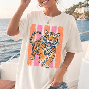 May include: An off-white short-sleeve t-shirt featuring a graphic print of a tiger with orange and black stripes. The tiger is positioned in front of vertical, distressed-style stripes in alternating shades of bright orange and hot pink.
