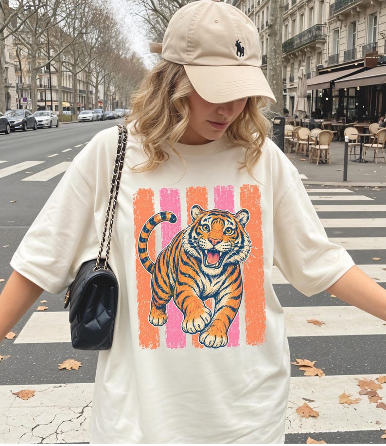 May include: A beige baseball cap with a small dark logo and an oversized cream-colored t-shirt featuring a graphic of a tiger in orange and black stripes against a background of vertical pink and orange textured stripes, paired with a black quilted bag.