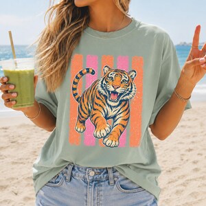 May include: A sage green short-sleeve t-shirt featuring a graphic print of a roaring tiger in orange and black. The tiger is positioned in front of four vertical stripes in alternating shades of pink and orange. The shirt has a crew neckline.
