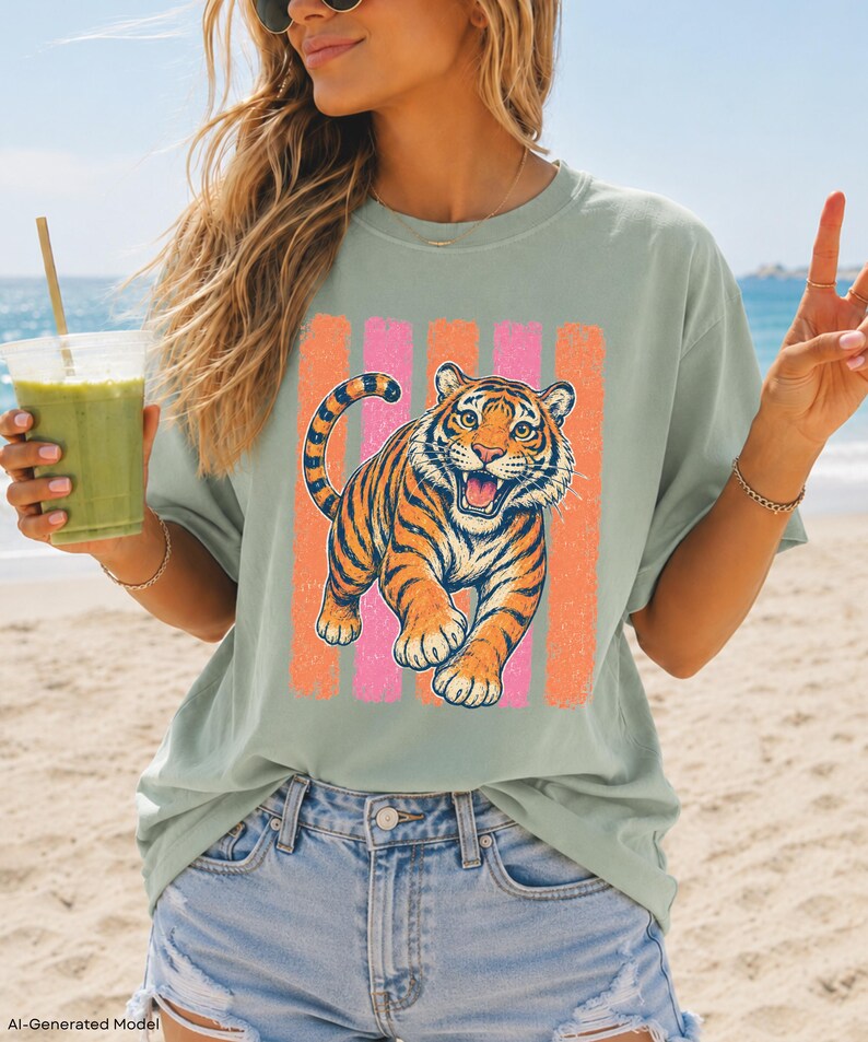May include: A sage green short-sleeve t-shirt featuring a graphic print of a roaring tiger in orange and black. The tiger is positioned in front of four vertical stripes in alternating shades of pink and orange. The shirt has a crew neckline.