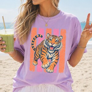 May include: A light purple short-sleeve t-shirt featuring a graphic print of an orange and black tiger in a pouncing position. The tiger is centered over a background of vertical, distressed-texture stripes in alternating shades of pink and orange.