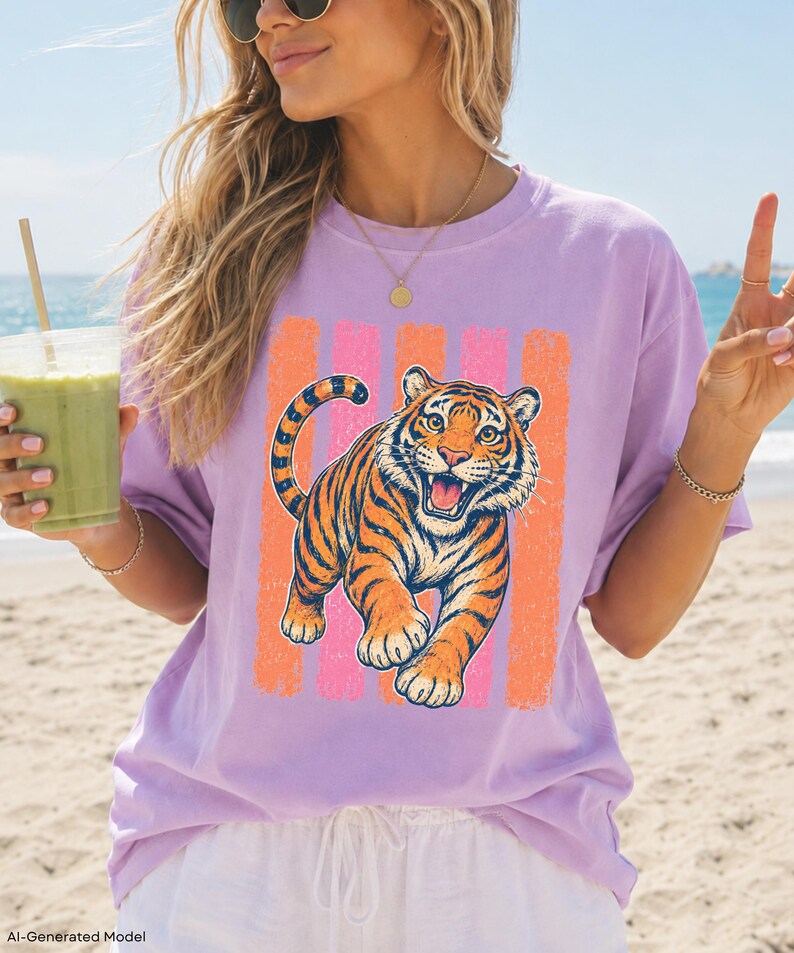 May include: A light purple short-sleeve t-shirt featuring a graphic print of an orange and black tiger in a pouncing position. The tiger is centered over a background of vertical, distressed-texture stripes in alternating shades of pink and orange.