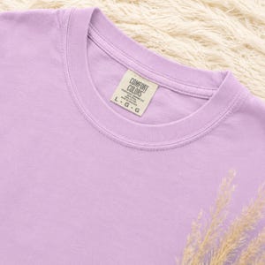 May include: A light purple crew neck t-shirt with a ribbed collar. The interior neck label reads COMFORT COLORS, 100% COTTON, MADE IN HONDURAS, and size L-G-G. The fabric has a solid, matte finish and a consistent pastel lavender hue throughout.