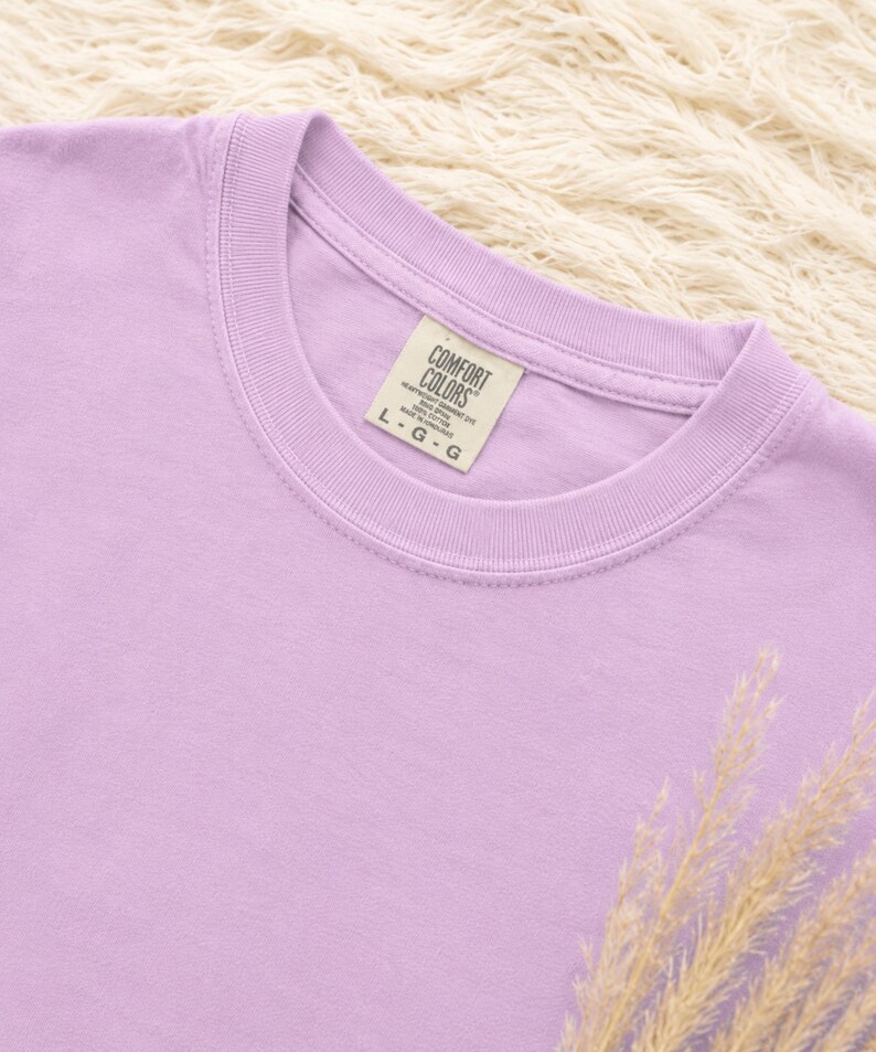 May include: A light purple crew neck t-shirt with a ribbed collar. The interior neck label reads COMFORT COLORS, 100% COTTON, MADE IN HONDURAS, and size L-G-G. The fabric has a solid, matte finish and a consistent pastel lavender hue throughout.