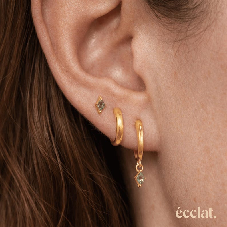 May include: Gold-toned earrings with a small green gemstone. The set includes a stud earring, a small hoop earring, and a larger hoop earring with a dangling gemstone. The word "&eacute;clat" is in the bottom right corner.