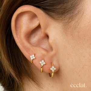 Puede incluir: Tres pendientes de aro dorados, cada uno adornado con un grupo de pequeñas piedras preciosas transparentes dispuestas en forma de flor de cuatro pétalos. Los pendientes se lucen en una oreja, mostrando su delicado diseño. La marca "éclat" es visible.
