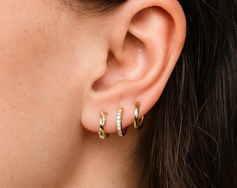 May include: A close-up of an ear adorned with three gold hoop earrings. The earrings vary in design, with one featuring a twisted rope pattern, another set with small, clear stones, and a classic plain hoop. The brand name "éccat." is visible.