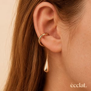 Könnte beinhalten: Ein goldener Ohrring mit einer glatten, polierten Schiene, die um den Knorpel verläuft, und einer feinen Goldkette, die nach unten hängt und in einem kleinen, glatten, tropfenförmigen Goldanhänger endet. Das Wort écclat. ist in Weiß gedruckt.