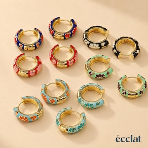 May include: A collection of eleven gold-tone hoop earrings featuring colorful enamel floral patterns in shades of blue, red, black, green, and orange. Each hoop has a hinged closure. The word "écclat" is printed in the bottom right corner.