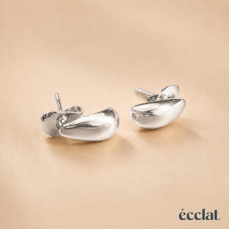 May include: A pair of shiny, silver-colored stud earrings. Each earring has a smooth, teardrop-like shape. The earrings are displayed on a light brown background. The brand name "&eacute;clat" is in the bottom right corner.