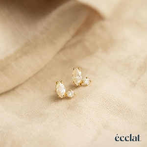 May include: A pair of gold stud earrings, each featuring a marquise-cut clear stone paired with a smaller round-cut clear stone. The earrings are set in a gold-tone metal. The word "écclat." is printed in the bottom right corner of the image.
