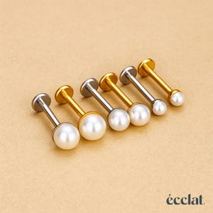 Puede incluir: Una selección de seis piercings labret con diseño de perla. Los piercings tienen un disco plano en un extremo y una perla en el otro. Algunos son dorados, otros plateados, cada uno con una perla en el extremo.