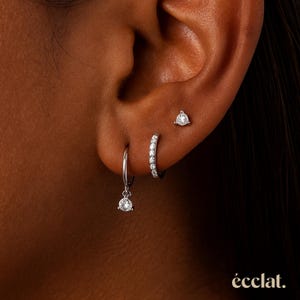 May include: A close-up of an ear adorned with three different silver earrings. One is a small stud, another is a small hoop with a dangling gem, and the third is a small hoop with gems along the front. The earrings are set against a dark background.