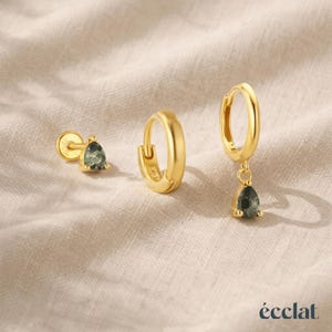 Może przedstawiać: Zestaw trzech złotych kolczyków. Jeden to sztyft z zielonym kamieniem w kształcie łzy. Drugi to mały kolczyk koła. Trzeci to kolczyk koła z zieloną zawieszką w kształcie łzy. Kolczyki są prezentowane na beżowej powierzchni.