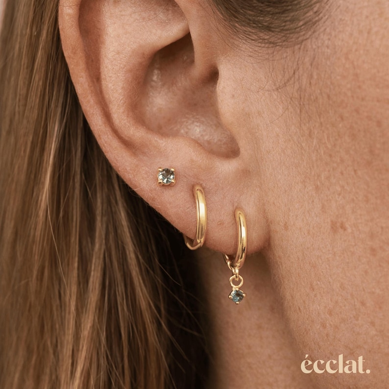 May include: A close-up of an ear adorned with three gold earrings. A small square stud earring is set in the upper ear. A gold hoop earring and a gold hoop earring with a small dangling square gemstone are also visible. The word "&eacute;clat." is at the bottom.
