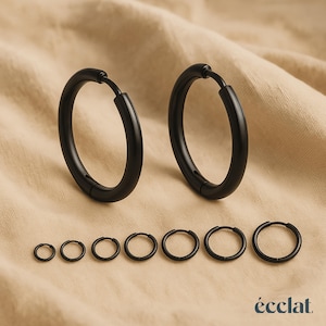 Può includere: Un set di orecchini a cerchio neri di varie dimensioni. L'immagine mostra due cerchi grandi e sei più piccoli. Gli orecchini sono realizzati in un materiale scuro e opaco e sono presentati su uno sfondo di tessuto neutro. Il marchio "éclat" è visibile.