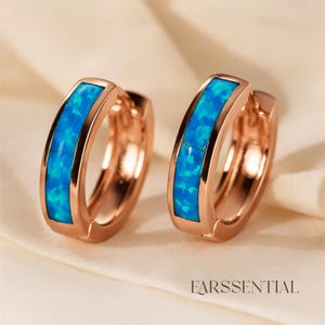 Peut inclure: Deux boucles d'oreilles cr&eacute;oles en or rose avec une incrustation d'opale bleu vif. Les boucles d'oreilles ont une finition polie et un fermoir s&eacute;curis&eacute;. Elles sont pr&eacute;sent&eacute;es sur une surface en satin blanc. Le mot "EARSSENTIAL" est imprim&eacute; en blanc en bas.