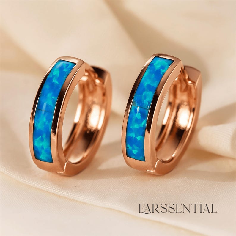 Peut inclure: Deux boucles d'oreilles cr&eacute;oles en or rose avec une incrustation d'opale bleu vif. Les boucles d'oreilles ont une finition polie et un fermoir s&eacute;curis&eacute;. Elles sont pr&eacute;sent&eacute;es sur une surface en satin blanc. Le mot "EARSSENTIAL" est imprim&eacute; en blanc en bas.