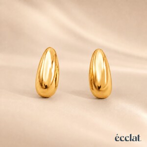 May include: Two polished gold teardrop-shaped earrings. The earrings are smooth and reflective, with a high-shine finish. The background is a soft, neutral color, and the brand name "&eacute;clat" is in the bottom right corner.