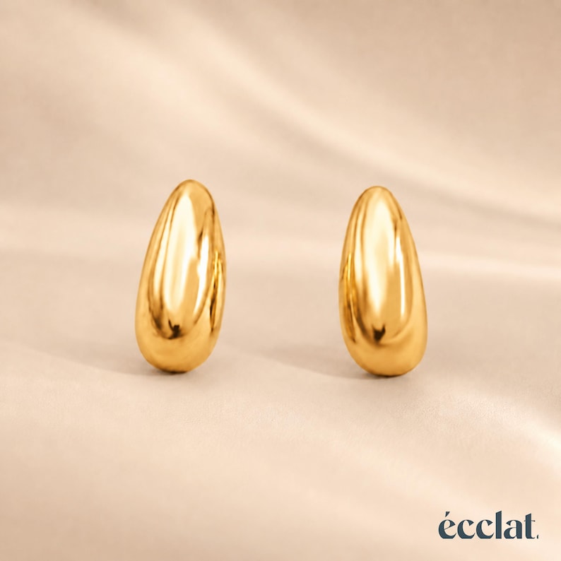 May include: Two polished gold teardrop-shaped earrings. The earrings are smooth and reflective, with a high-shine finish. The background is a soft, neutral color, and the brand name "&eacute;clat" is in the bottom right corner.