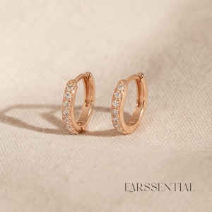 May include: Two rose gold hoop earrings, each featuring a row of small, clear gemstones. The earrings are presented against a neutral backdrop. The word "EARSSENTIAL" is printed in a serif font at the bottom of the image.
