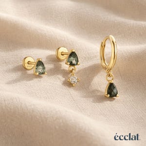 May include: A selection of gold earrings with green, teardrop-shaped gemstones. The set includes a stud earring, a dangle earring with a small diamond-like accent, and a hoop earring. The earrings are displayed on a beige fabric surface.