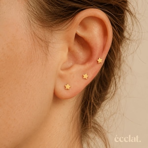 May include: Three small, gold-colored star-shaped stud earrings are positioned along the earlobe and cartilage. Each earring features a simple, five-pointed star design with a smooth, polished finish, creating a uniform look across the ear.