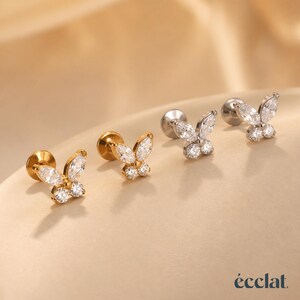 May include: Four butterfly-shaped stud earrings. Two are gold-toned, and two are silver-toned. Each earring features a butterfly design with clear, sparkling crystals. The earrings are displayed on a neutral, light-colored surface.